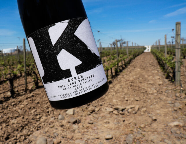 A bottle of Phil Lane Syrah wine in front of grapevines growing in the background and a large letter 'K'